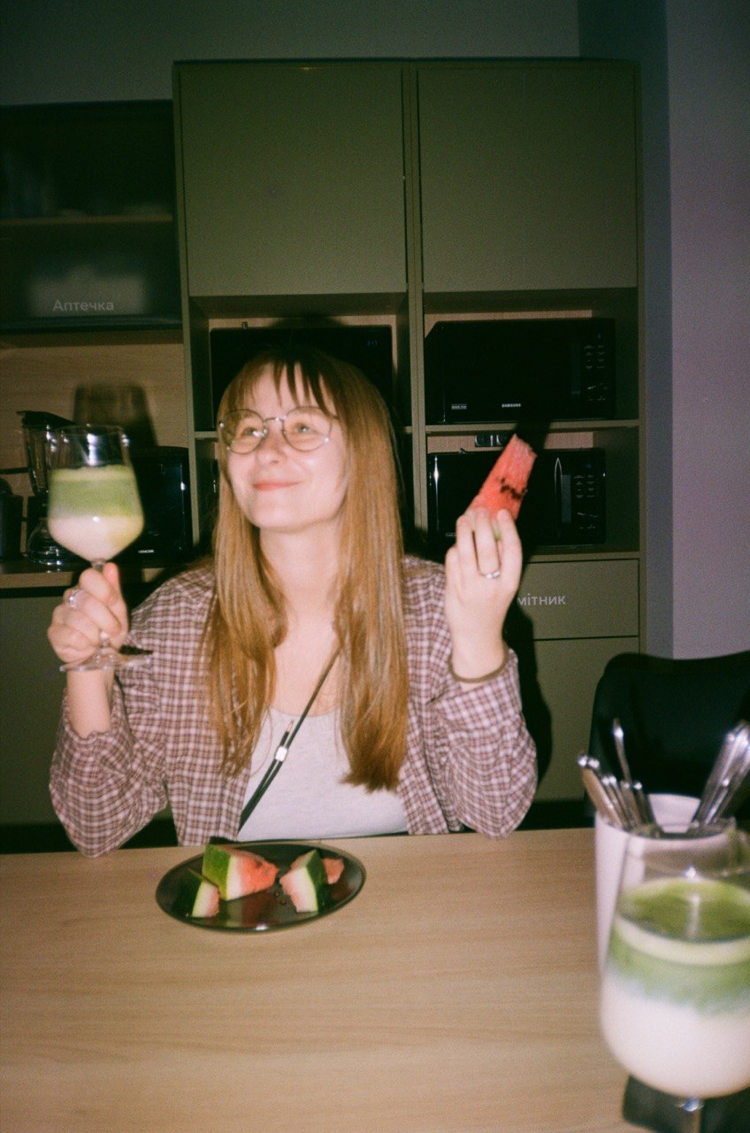 Anastasiia smiling at a table, holding a glass and a slice of watermelon
